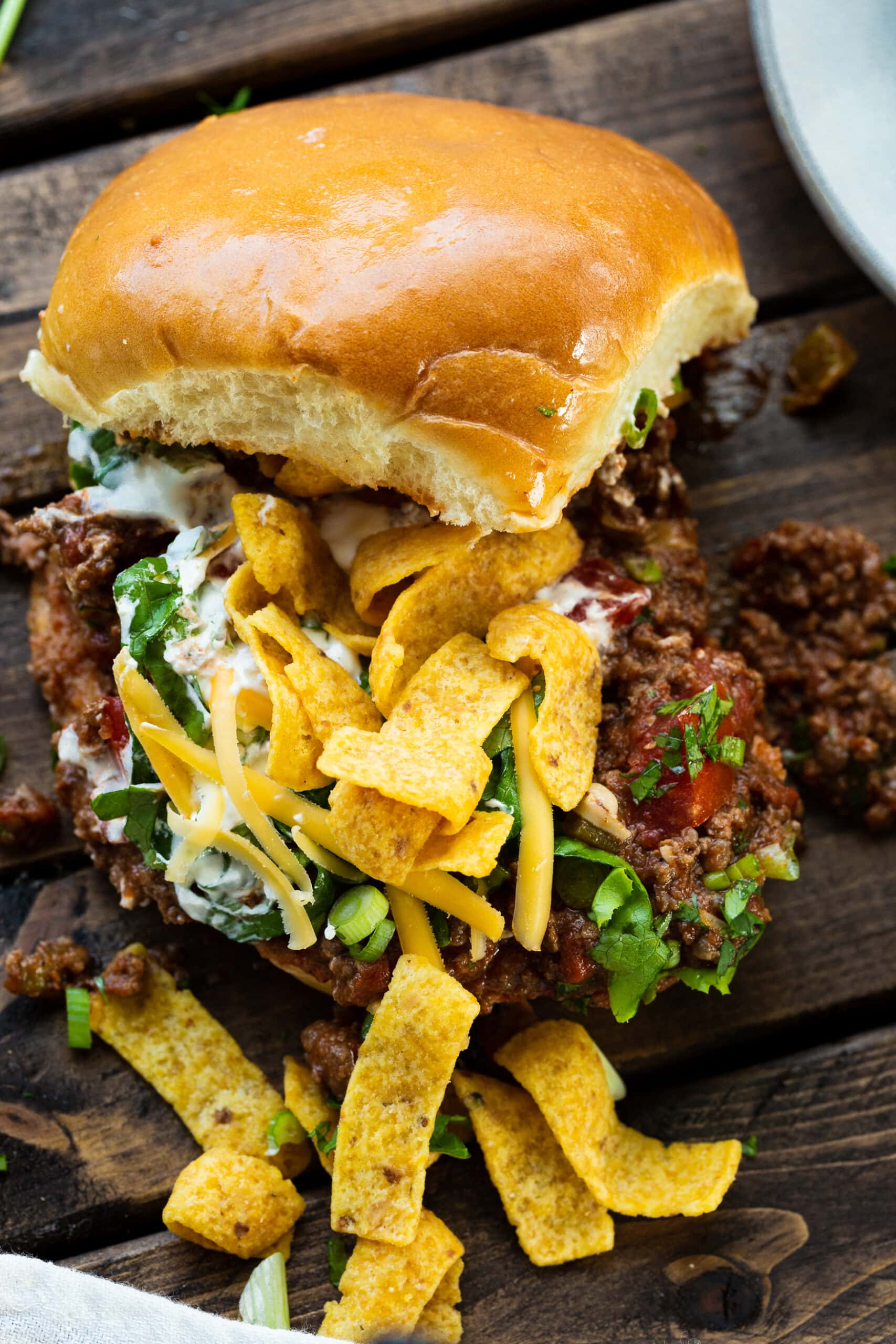 a photo of a hamburger bun piled with ground beef taco meat topped with shredded lettuce, Fritos, sour cream, diced tomatoes and cheddar cheese with the top bun sitting on top.