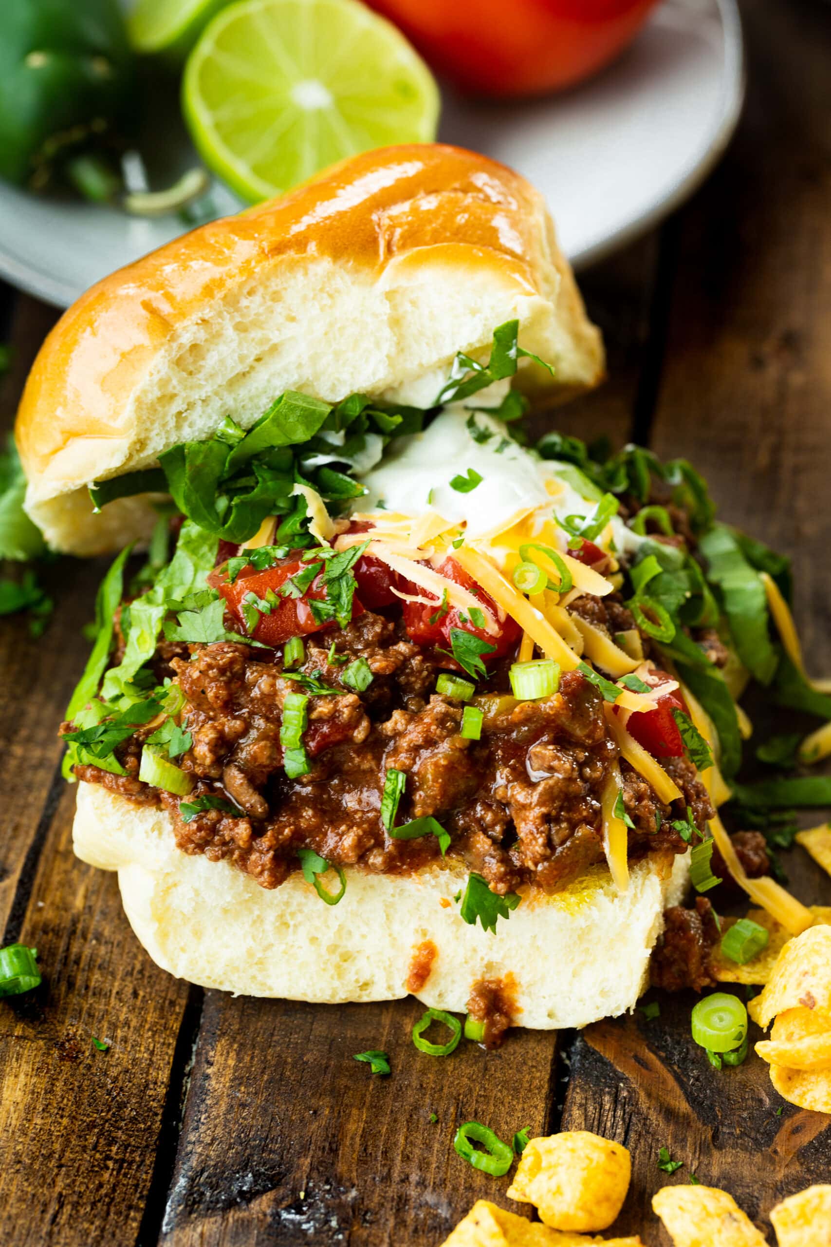 a photo of a brioche hamburger bun loaded with taco meat topped with shredded lettuce, cheddar cheese, diced tomatoes, sour cream and fritos chips scattered around.