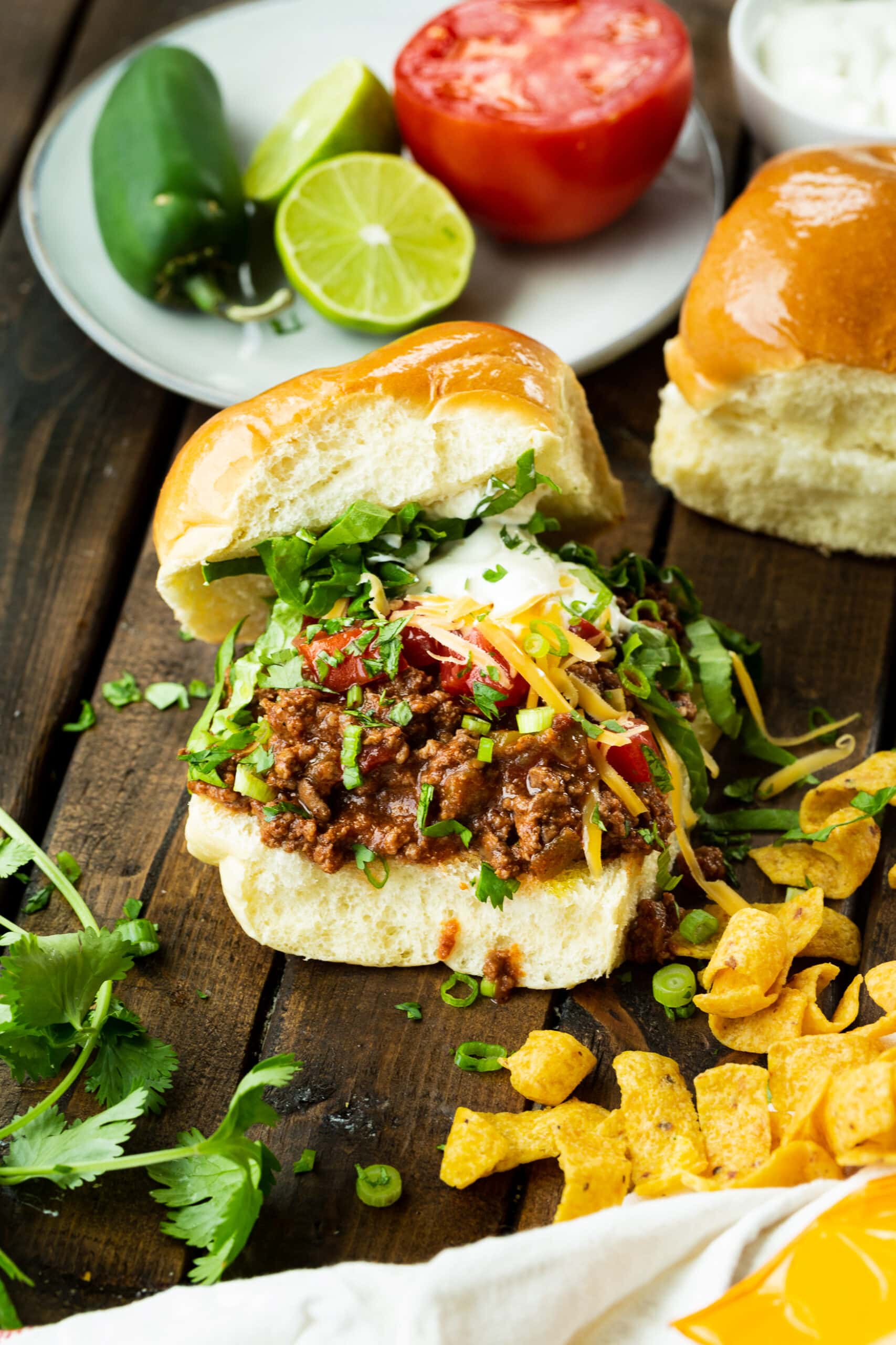 a photo of a hamburger bun piled high with juicy taco meat topped with shredded lettuce, sour cream, shredded cheddar cheese, sliced green onions and diced tomatoes with Fritos scattered around the sandwich.