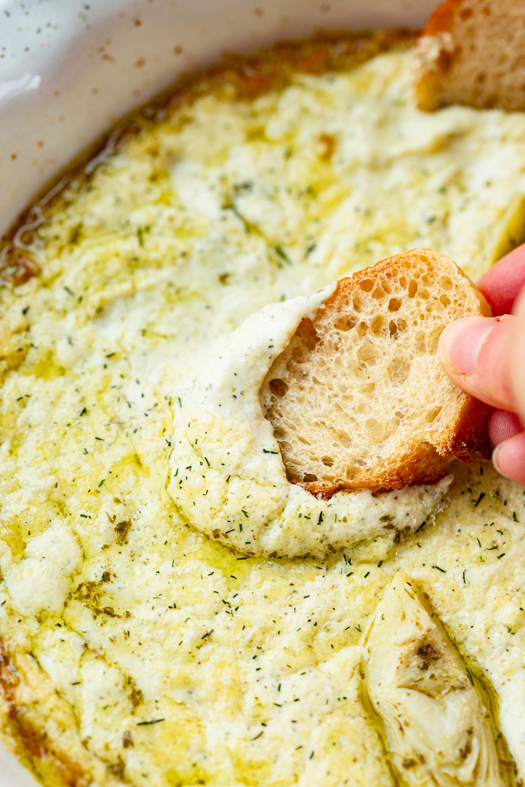 a photo of a hand dipping a crostini in a large dish filled with creamy artichoke dip.
