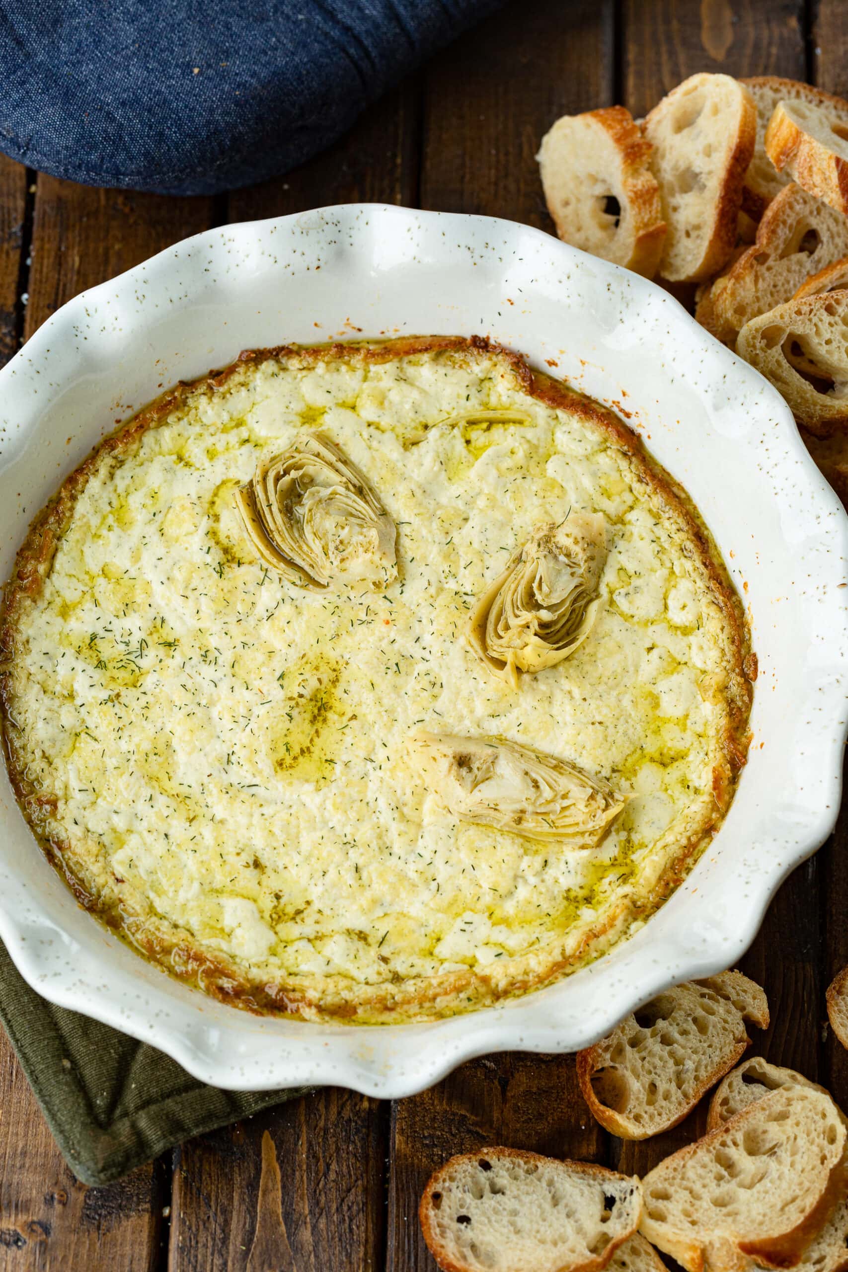 a photo of a white ceramic circular baking dish full of golden creamy artichoke dip with several pieces of artichoke placed on top.