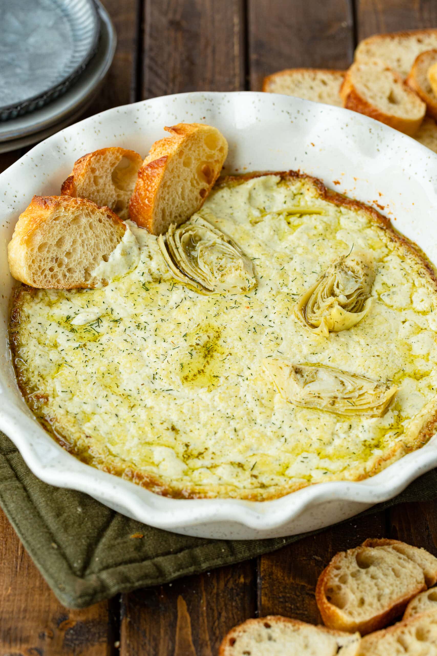 a photo of a circular white ceramic baking dish full of golden creamy artichoke dip with three slices of baguette stuck in the dip along the side of the pan.