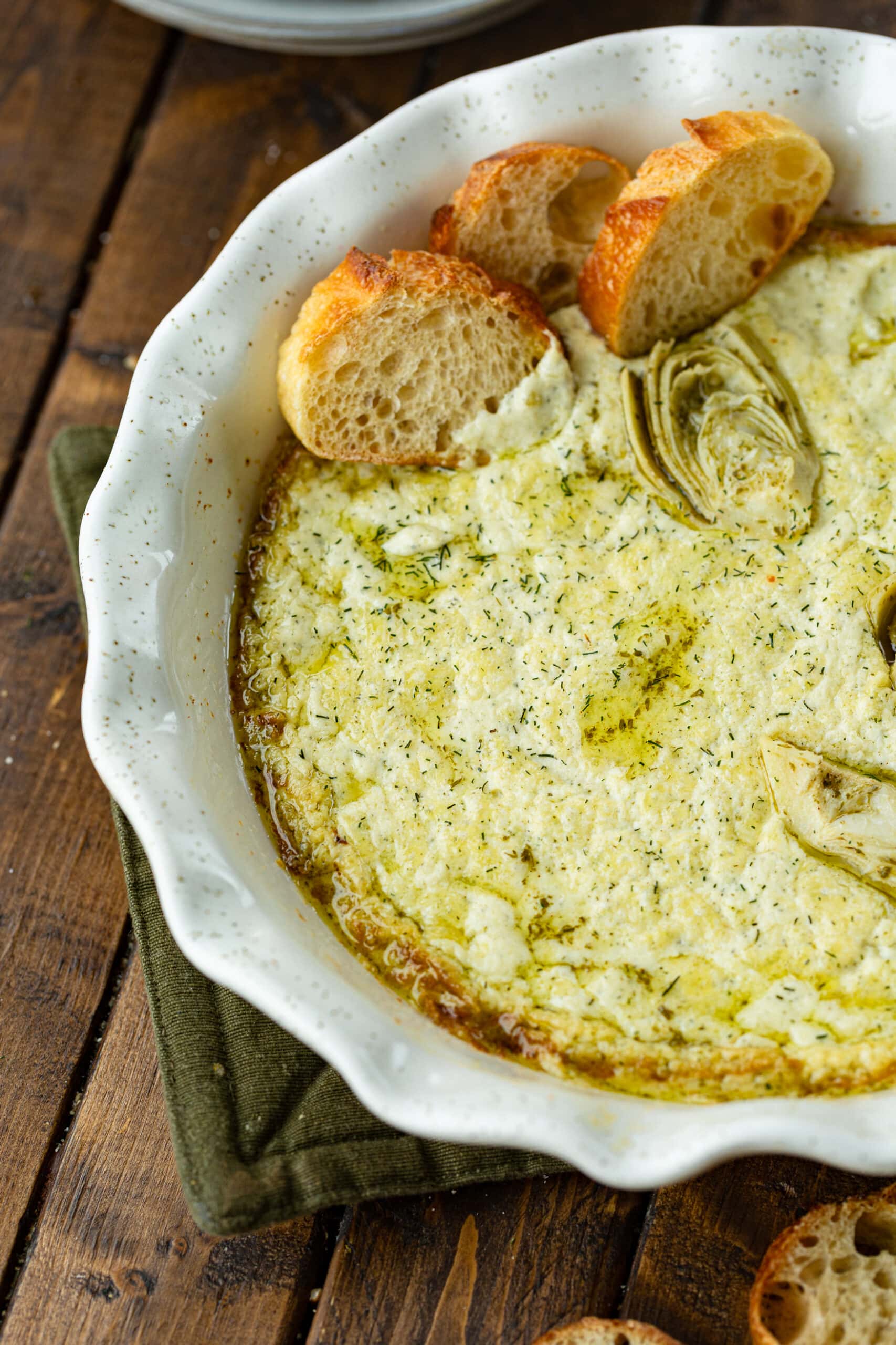 a close up photo of a baking dish full of baked creamy artichoke dip topped with chunks of artichoke and slices of baguette.