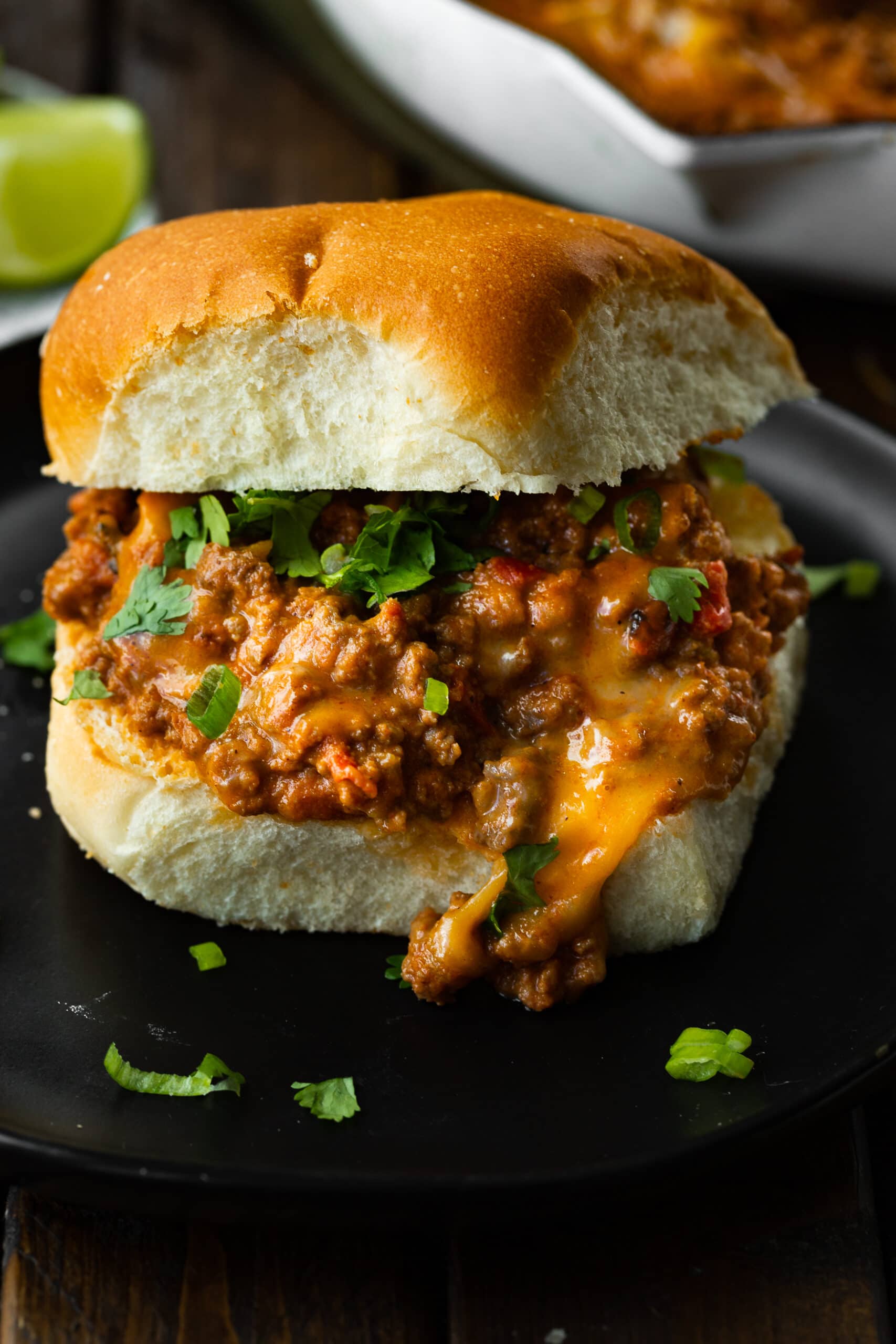 a photo of a cheesy taco sloppy joe with the top bun on top and chopped cilantro and scallions sprinkled on top.